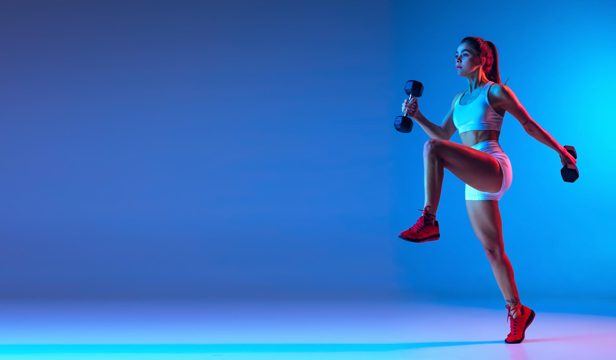 Portrait of young sportive girl training with dumbbells isolated over blue background in neon