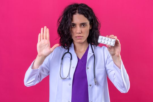 Woman doctor wearing white coat with stethoscope standing with open hand making stop gesture and holding blister of pills in other hand on isolated pink
