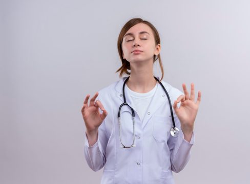 Peaceful young female doctor wearing medical robe and stethoscope doing ok sign with closed eyes  with copy space