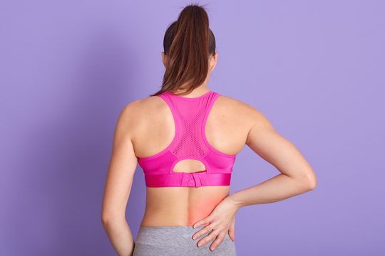 Back view of young woman with back pain standing against purple wall in studio