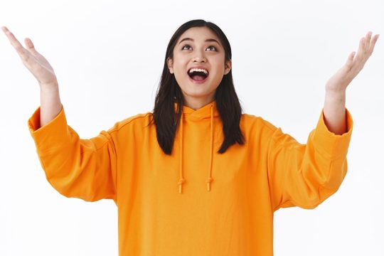 Amused and happy triumphing young asian girl in orange hoodie raising hands up sky and smiling looking up thankful catching something or thanking heaven for dream come true