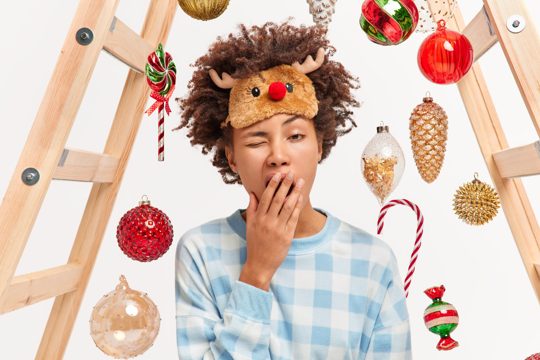 Photo of exhausted woman wakes up very early in morning yawns and wants to sleep has much to do before new year dressed in checkered pajama and sleep mask decorates house with christmas baubles