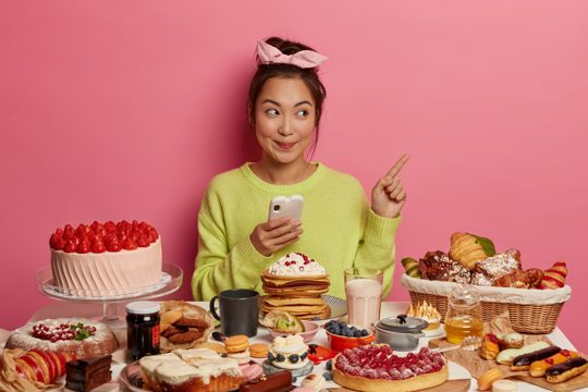 Cheerful glad brunette asian girl surrounded by biscuits, cookies and cakes, enjoys sweet food during festive time, enjoys holidays treats, points at blank space, uses mobile phone.