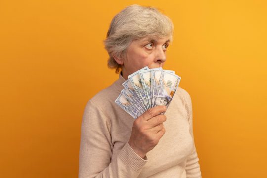 Old woman wearing creamy turtleneck sweater standing in profile view holding money looking up from behind it isolated on orange wall with copy space