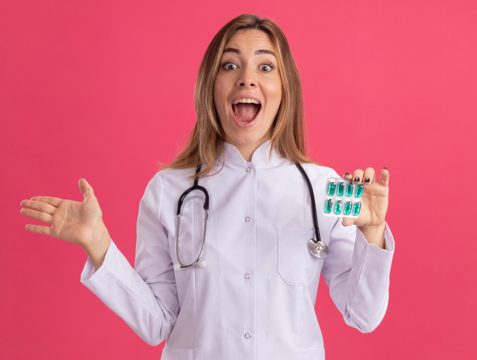 Surprised young female doctor wearing medical robe with stethoscope holding pills spreading hand isolated on pink wall