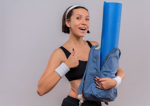 Young fitness woman in sportswear holding backpack with yoga mat with smile on face showing thumbs up standing over white wall